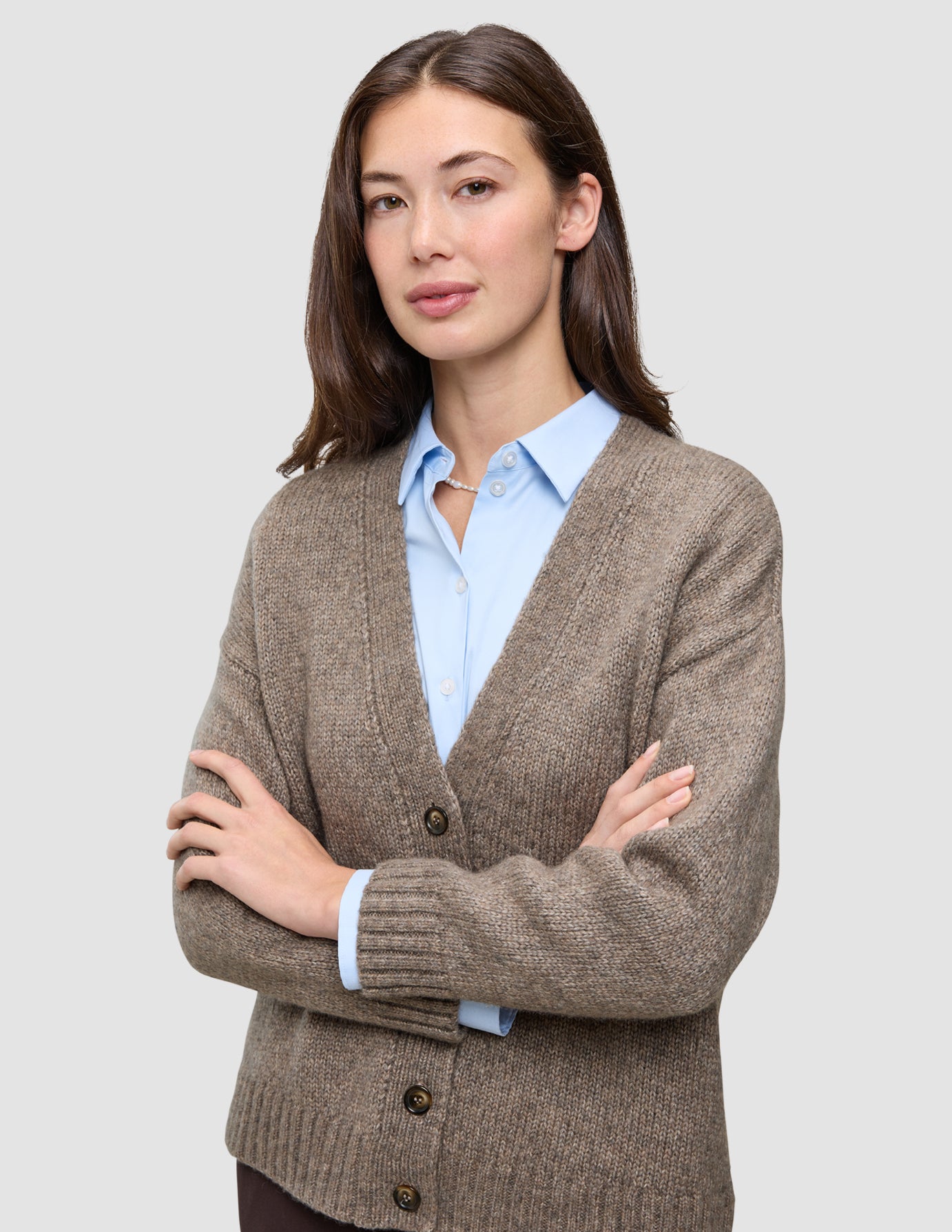 En kvinde iført en tyk, blød uldstrikket cardigan i farven taupe melange over en lyseblå skjorte med knapper.